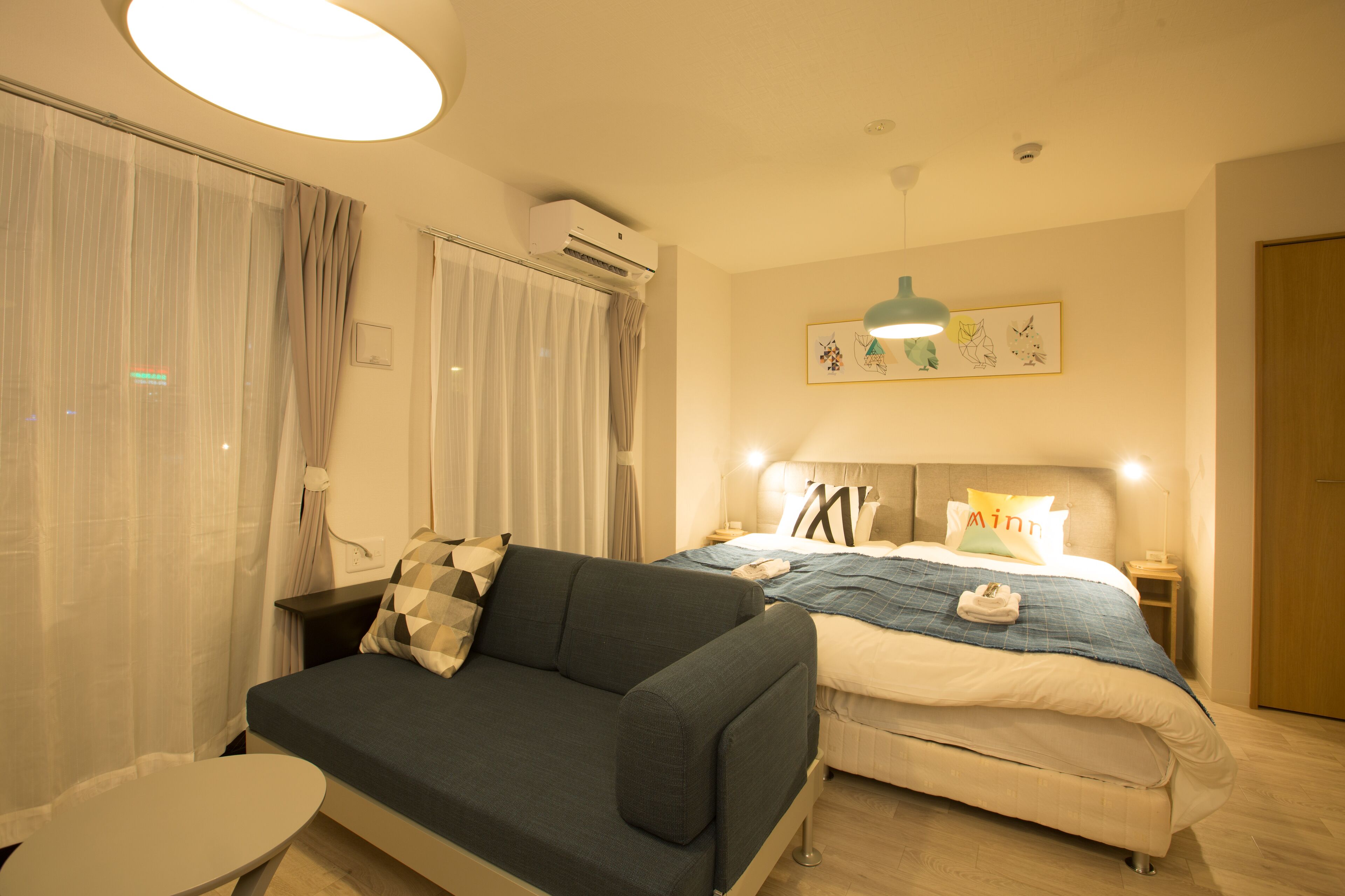 A modern hotel room with a sofa, a bed, a small table, and a ceiling-mounted air conditioner.