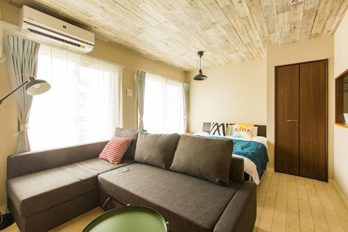 A room with a sectional sofa, a bed with a blue blanket, a wooden wardrobe, and an air conditioning unit.
