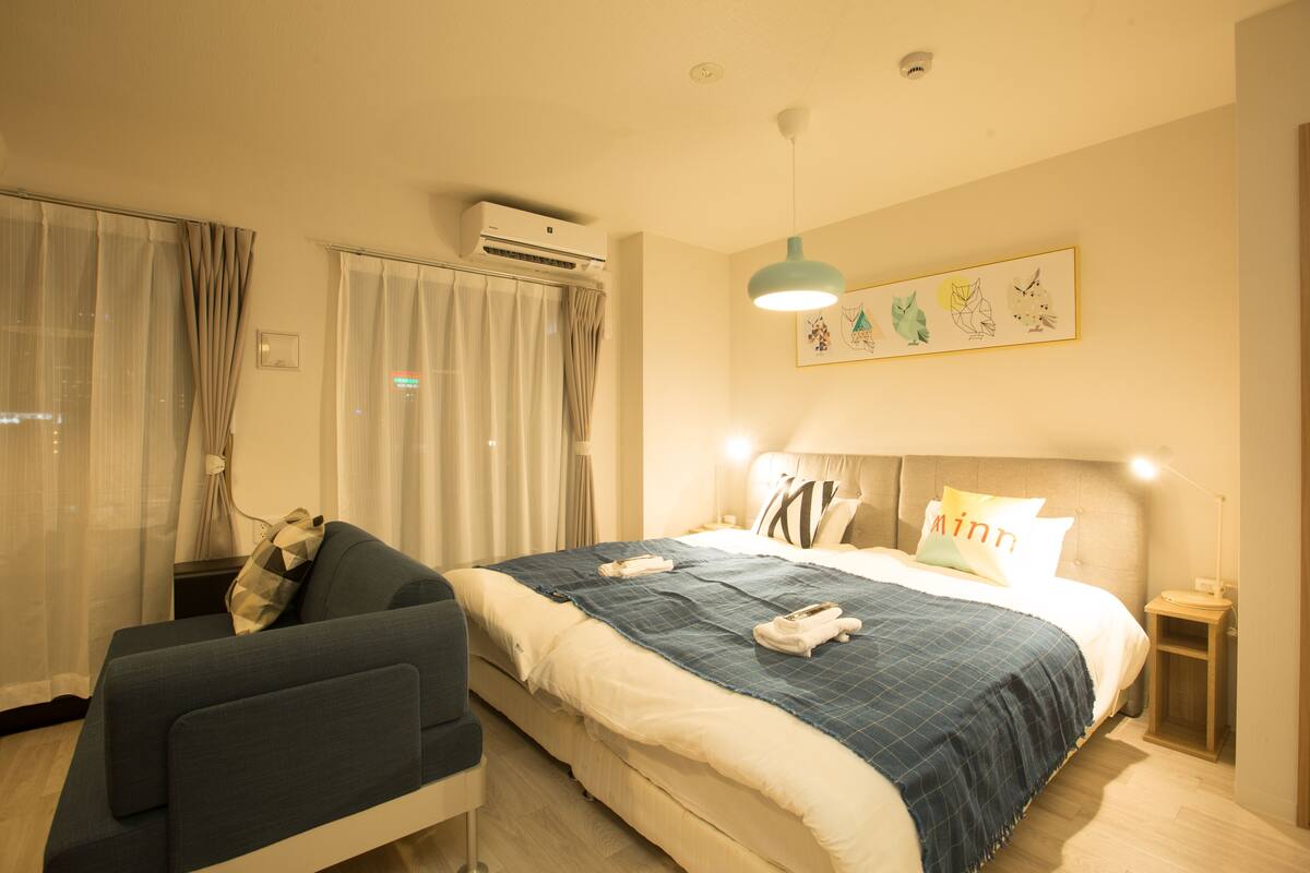 A hotel room with a bed, a chair, an air conditioning unit, a wall-mounted artwork, and a bedside table with a lamp.