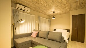 A modern, well-lit room with a sectional sofa, a small round table, and a bed with a wooden headboard. - Skyliving Namba (Osaka)