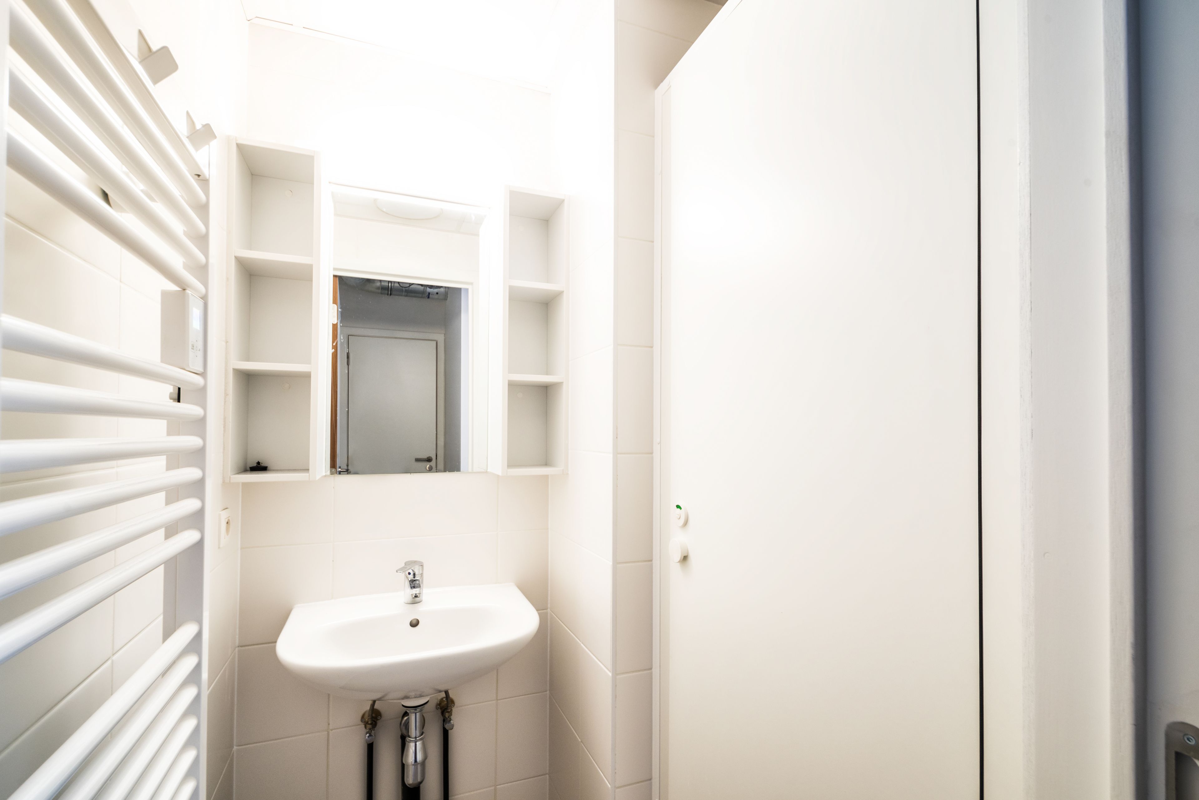 Un baño con lavamanos blanco, espejo y estantes abiertos.