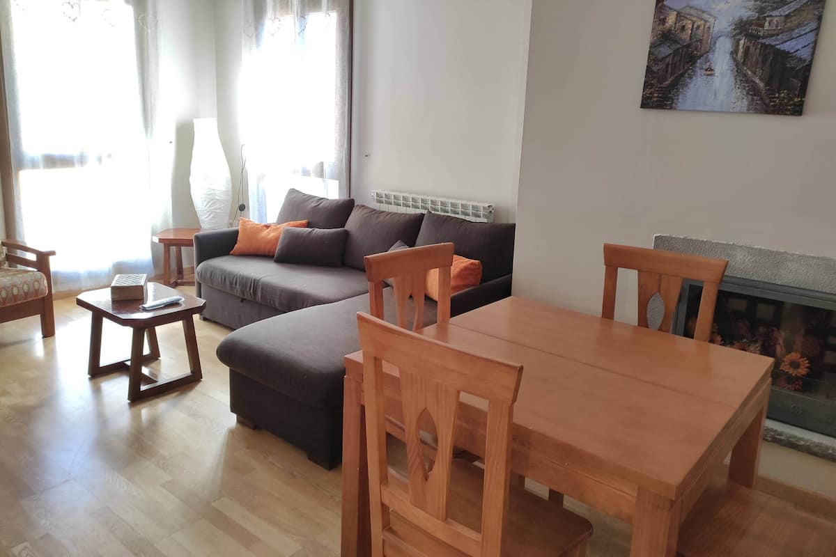 A living room with a sofa, ottoman, wooden table, chairs, and a painting on the wall.