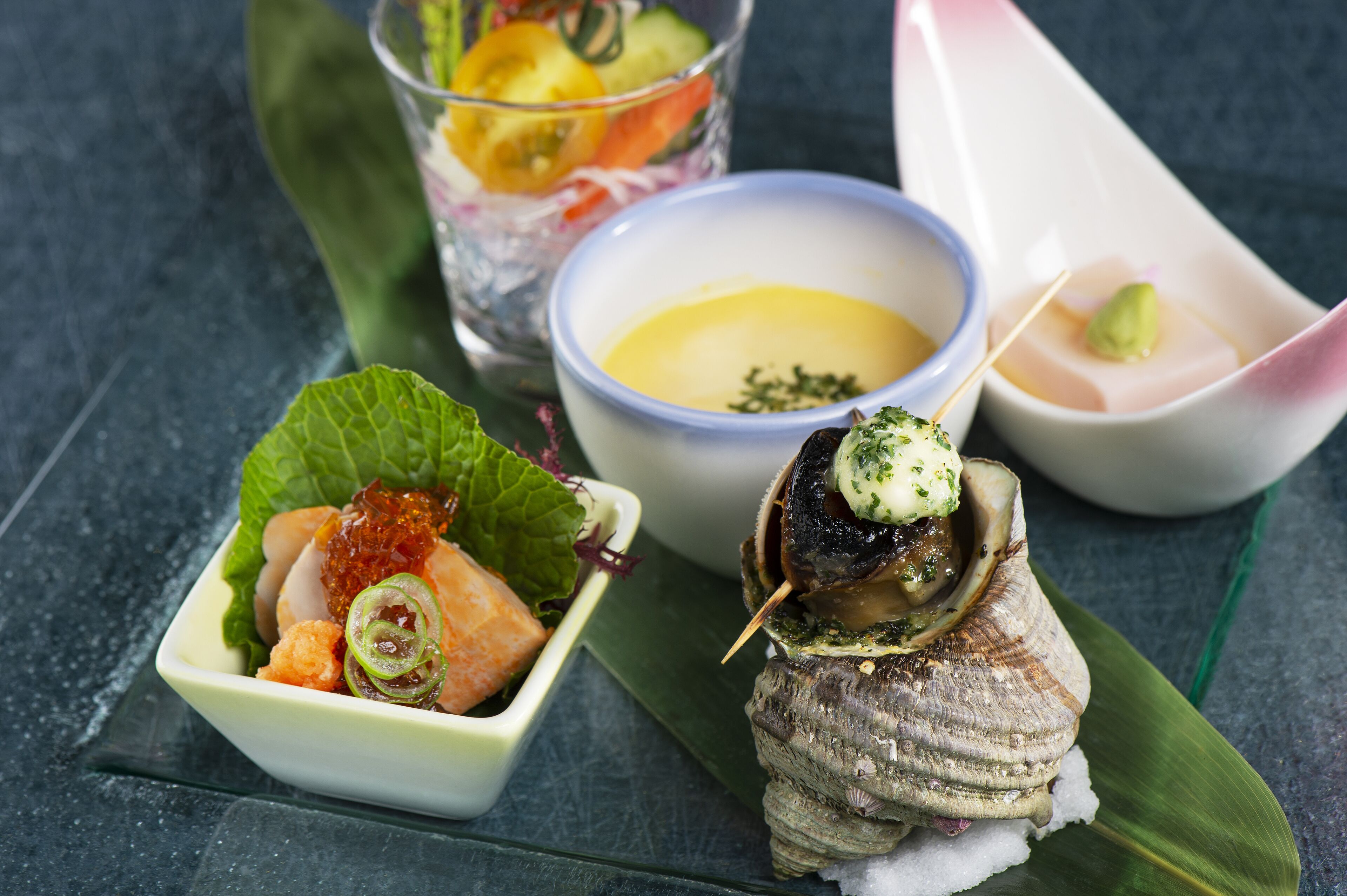 A seafood dish with a bowl of sauce, a glass of salad, and a plate with a shell.