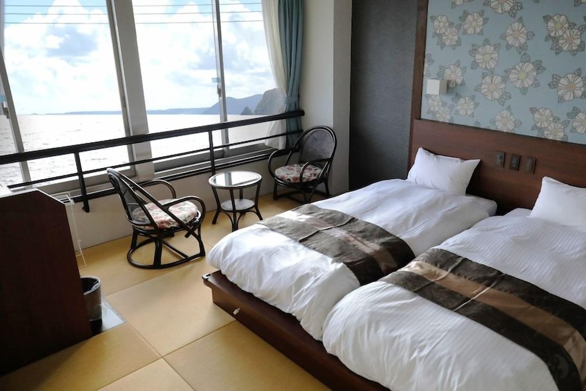 A hotel room with a large bed, two chairs, a small table, and a view of the sea through large windows.