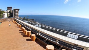 A wooden deck with a railing overlooking the sea, a building with multiple balconies, and a clear sky. - RYUGU no TSUKAI (Higashiizu)