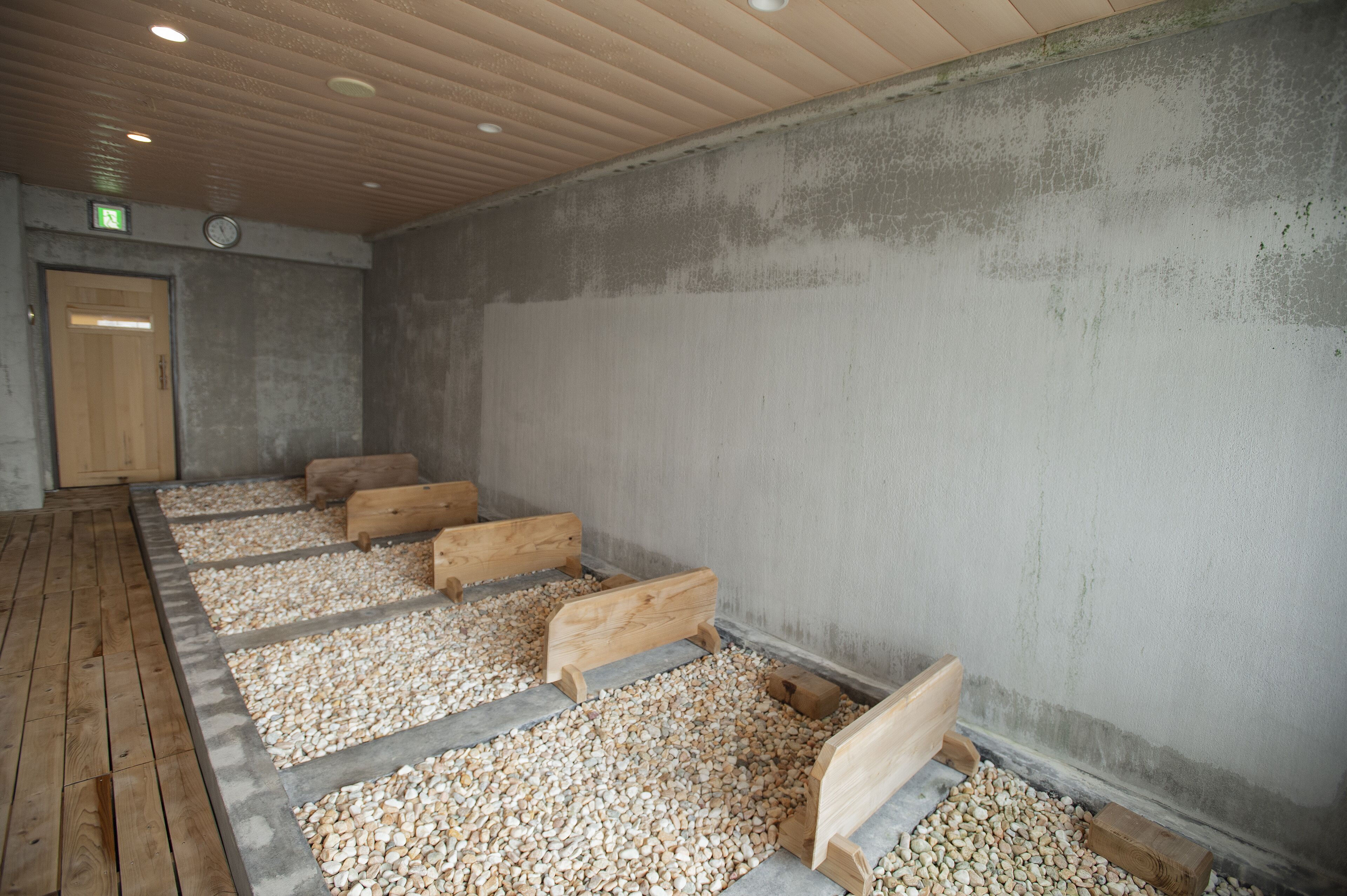Ein Raum mit an einer Wand entlang gereihten Holzbänken, Holzdielenboden und einer Betonwand mit einer Uhr über einer Tür.