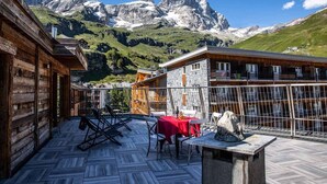 Eine Holzdiele mit Tisch und Stühlen, mit Blick auf ein Gebirge.