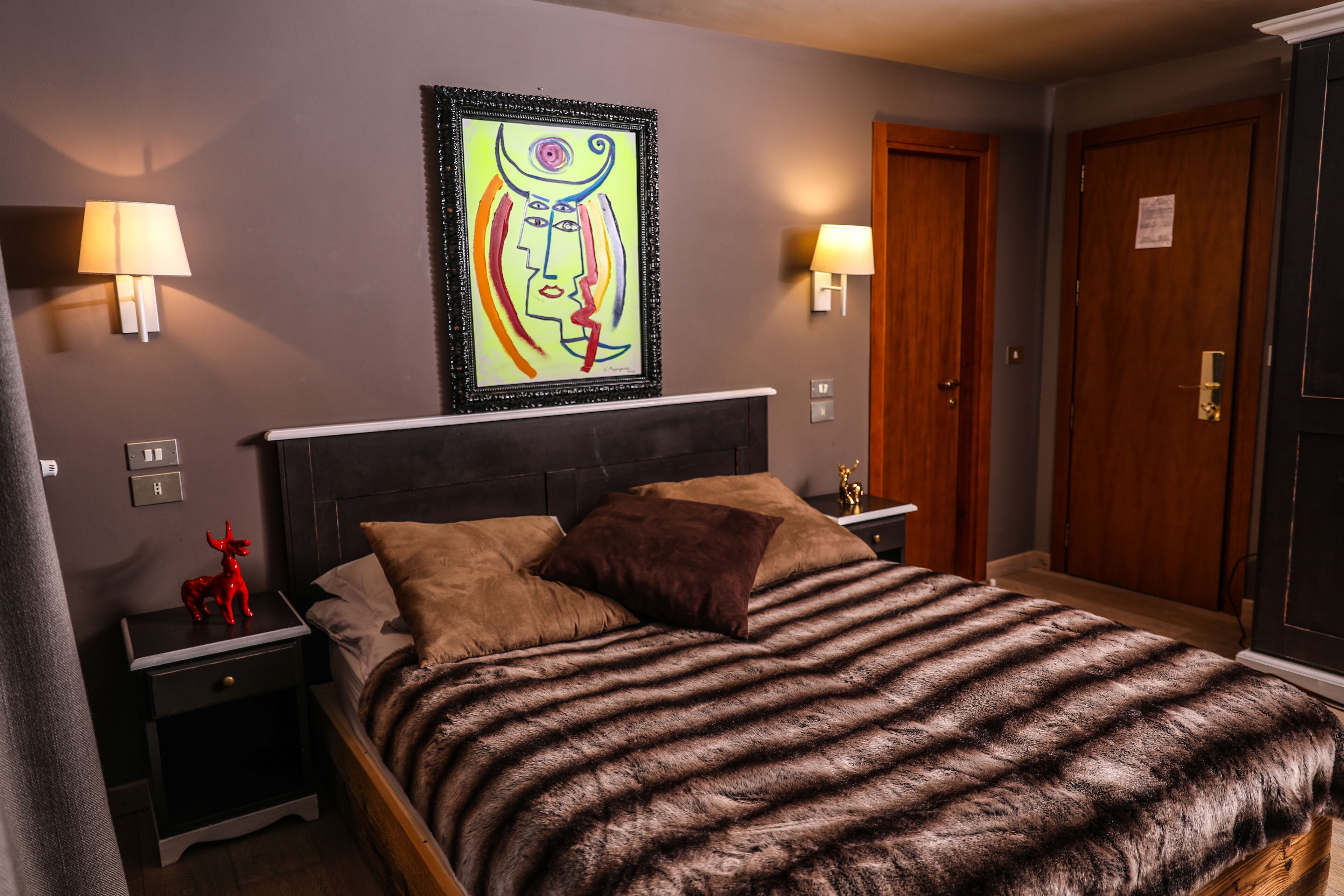 A bedroom with a large bed, two nightstands, a red statue, a framed abstract painting, and a door.