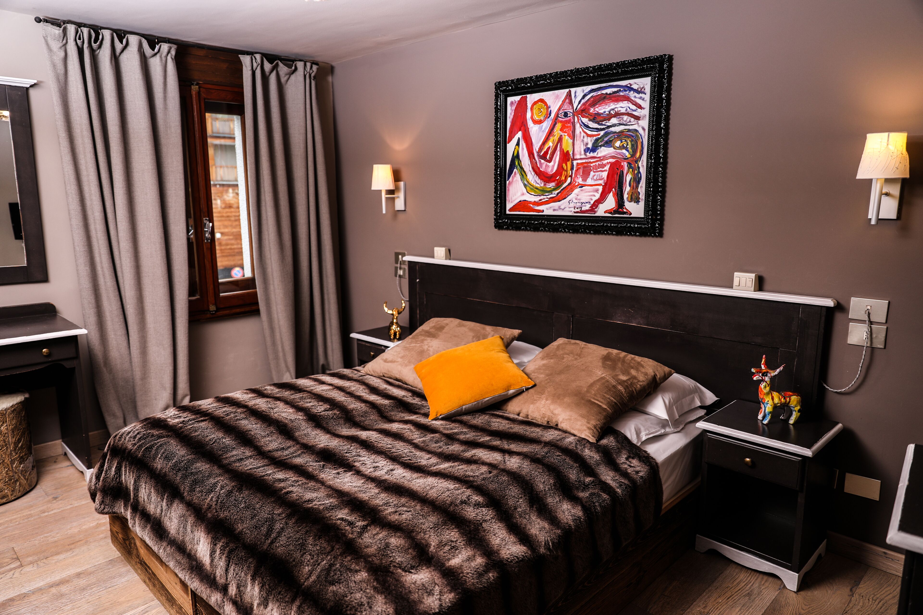 A bedroom with a large bed, two bedside tables, a nightstand, a desk, a chair, a window with curtains, and a wall-mounted painting.