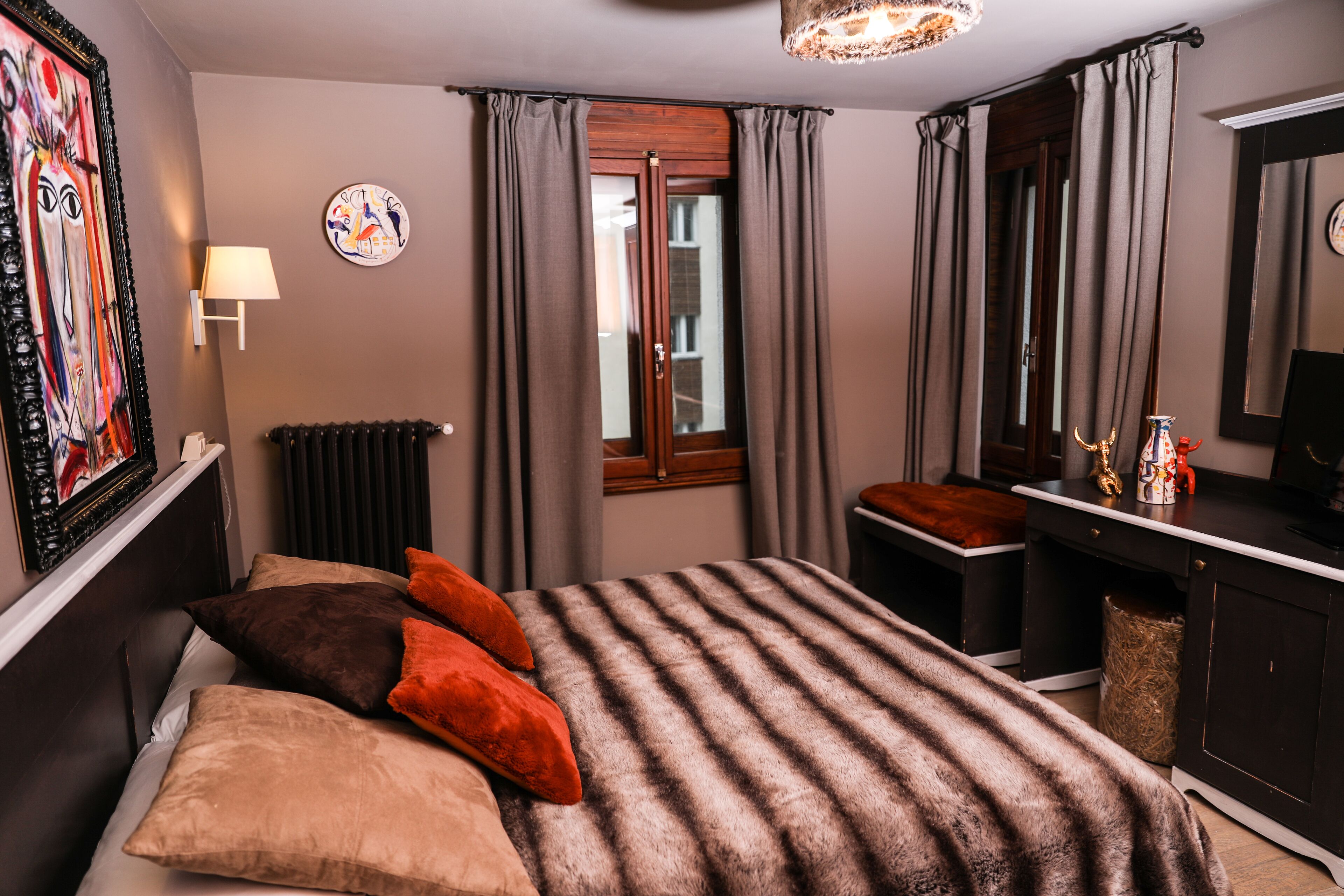 A bedroom with a bed, a desk, a chair, a radiator, a window, and a painting on the wall.