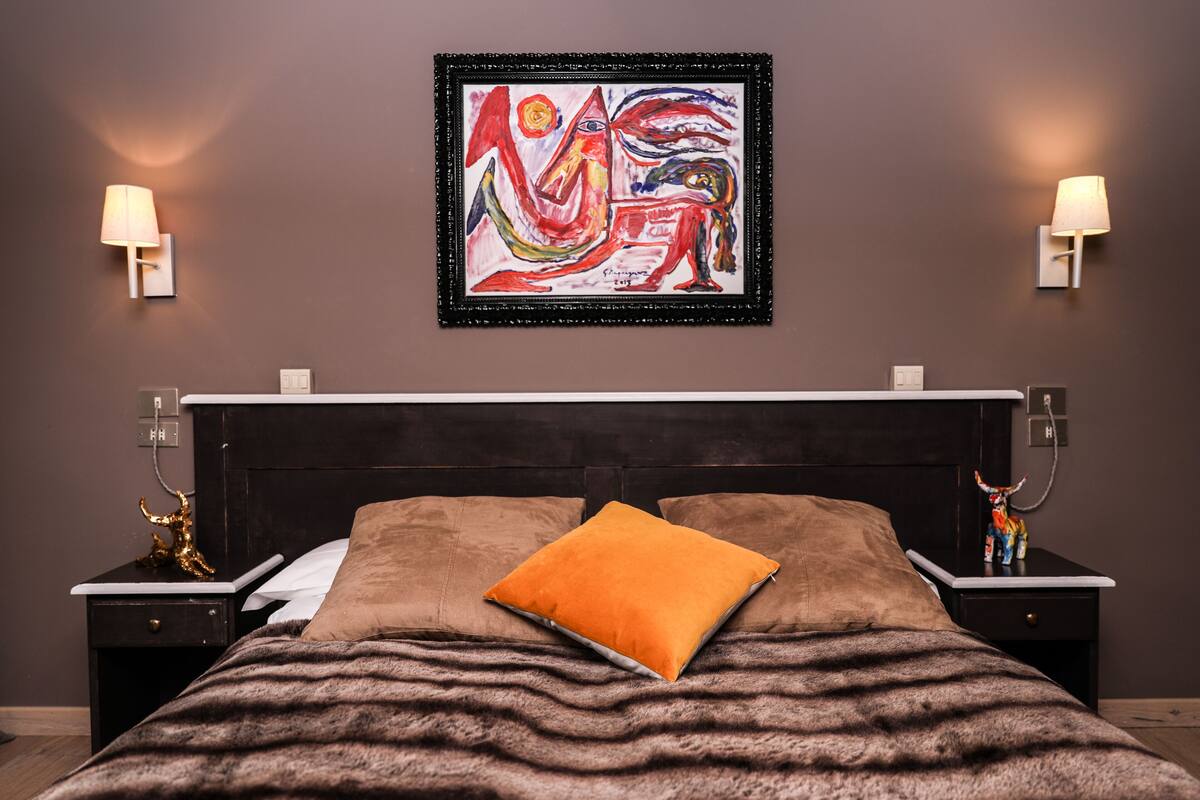 Une chambre à coucher avec une tête de lit sombre, une couverture à motifs zébrés, un coussin orange et un tableau abstrait coloré accroché au mur.