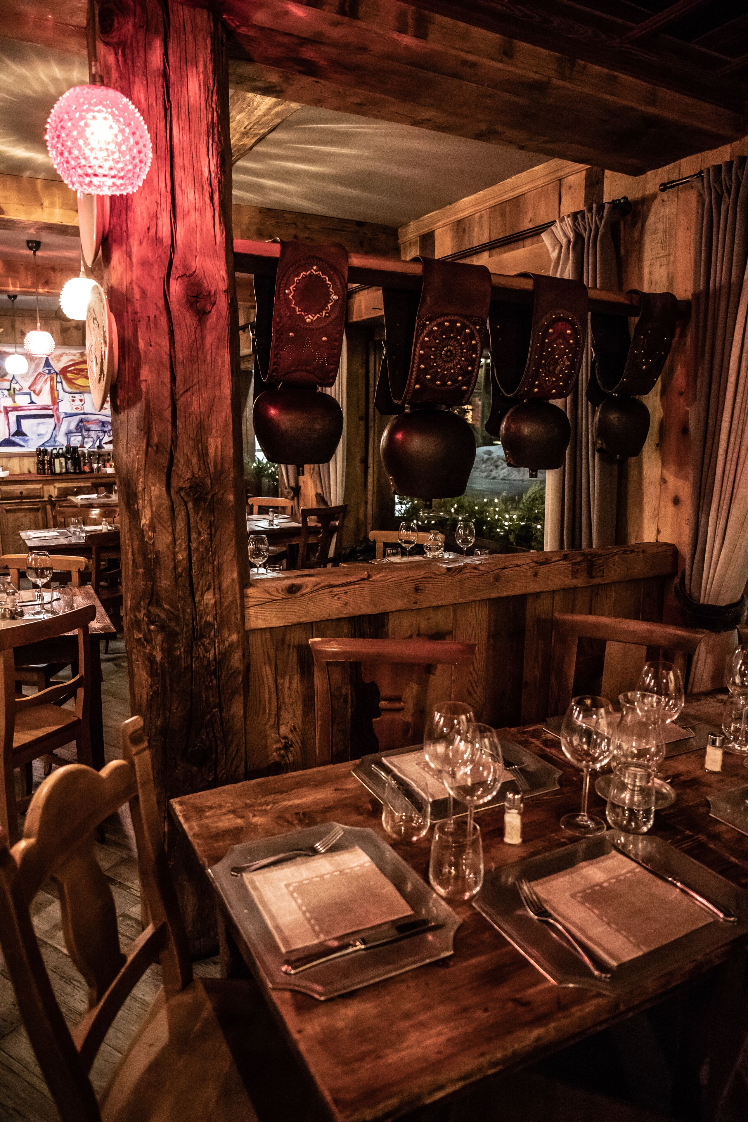 A rustic restaurant interior with wooden tables set for dining, hanging lanterns, and a view of greenery outside.