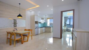 A modern kitchen with white cabinets, a dining area with wooden chairs, and a view through an open door to a balcony. - Beach City Apartment (Bac Nha Trang)
