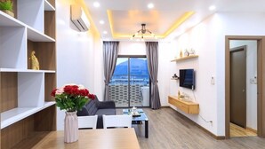A modern living room with a dining area, a large window with curtains, a mounted TV, and a view of the cityscape. - Beach City Apartment (Bac Nha Trang)