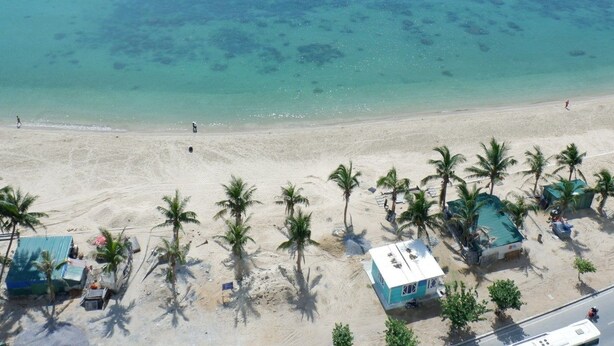 A beach with clear turquoise water, several boats, and a few people. There are palm trees and a small building with a blue roof. - Beach City Apartment (Bac Nha Trang)