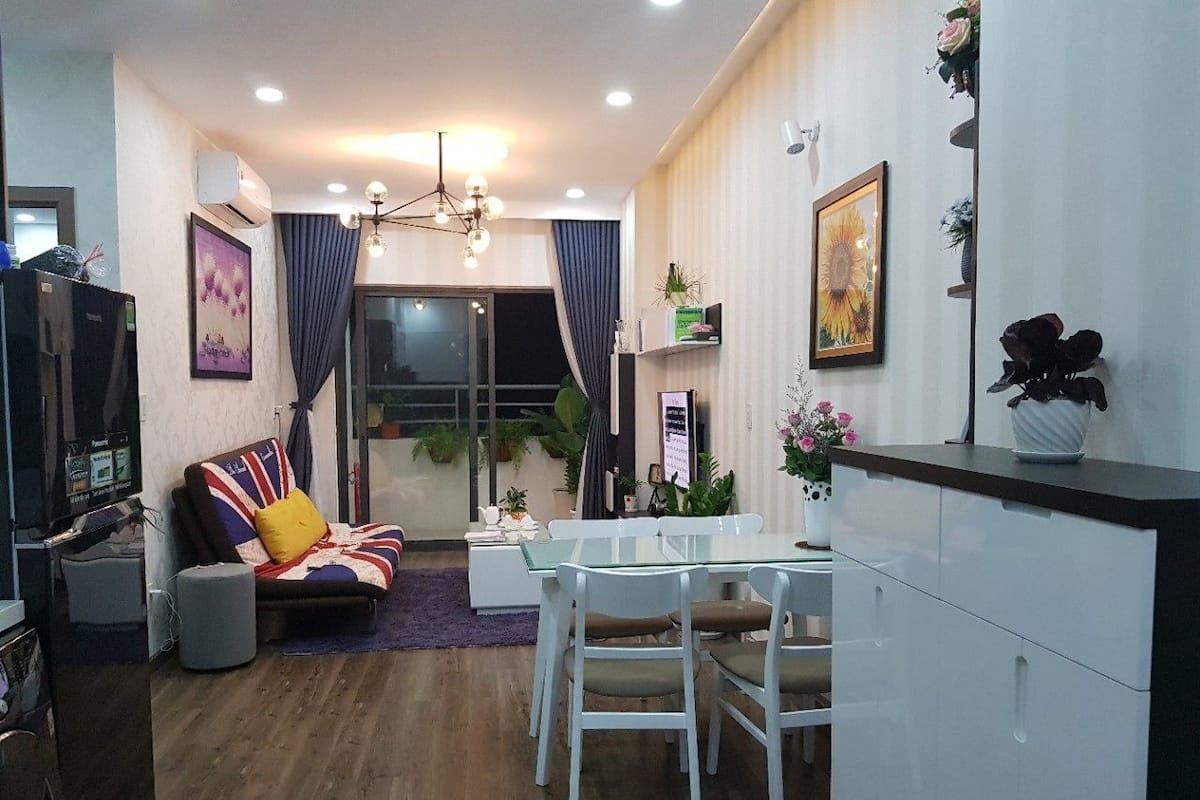A modern living room with a sofa, dining table, and chairs. There is a TV, a ceiling fan, and a window with curtains.
