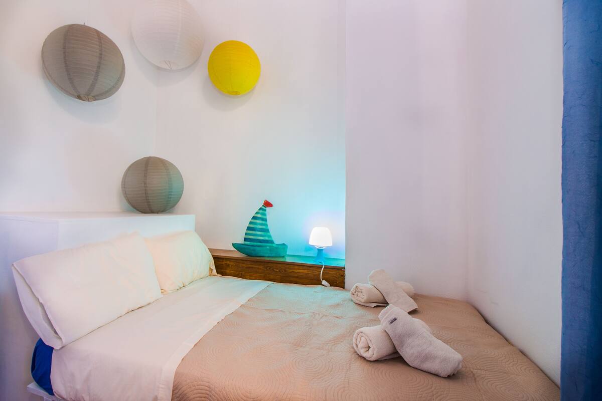 A neatly made bed with pillows and a blanket, a small bedside table with a lamp and a decorative ship model, and three wall-mounted spherical objects.