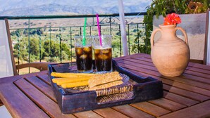 A table with a tray of food, a vase with flowers, and two drinks under an umbrella. - Malaxiana Houses (Chania)