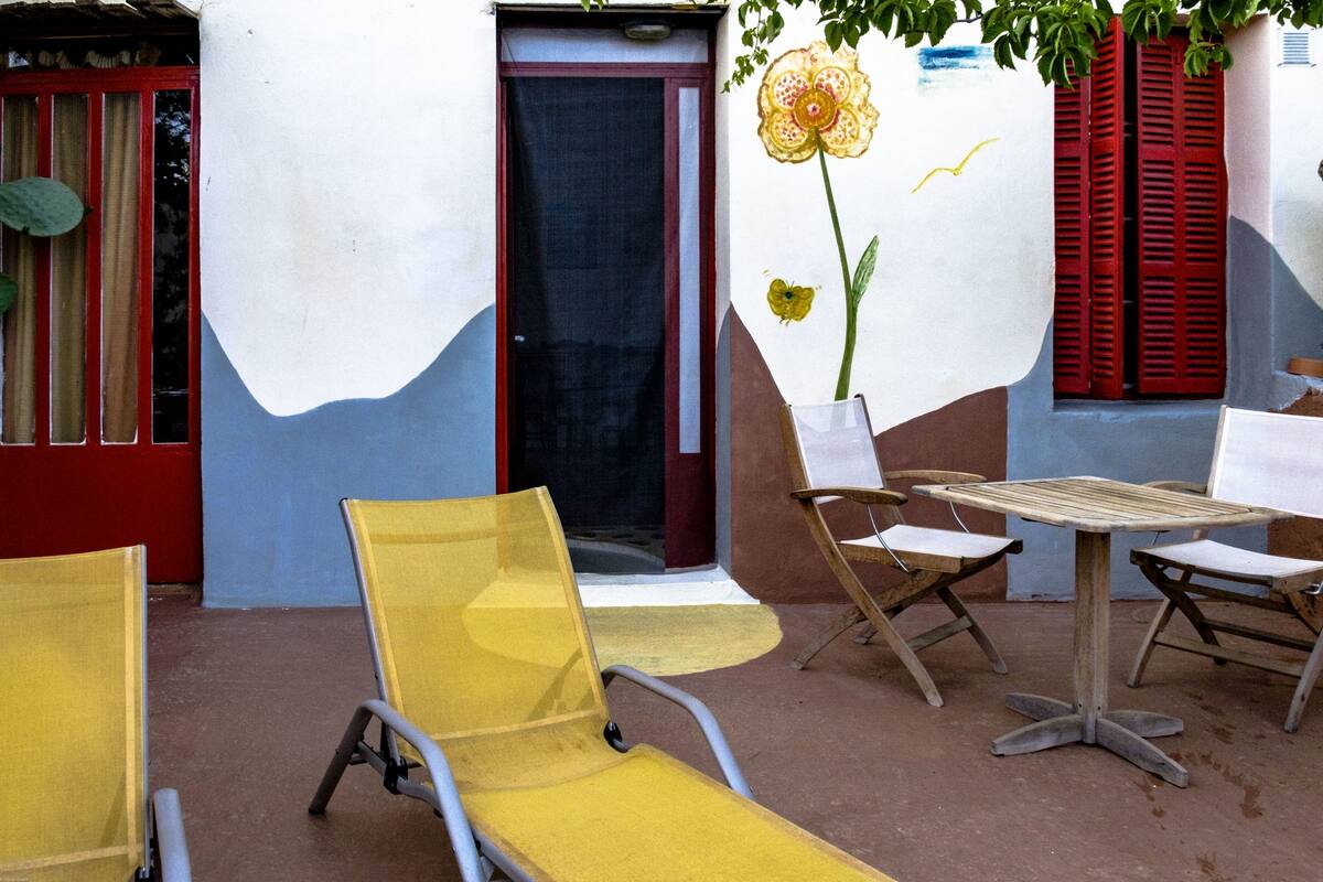 A patio with yellow chairs and a wooden table, a red door, and a wall with a flower painted on it.