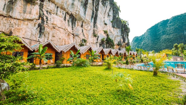 A row of thatched-roof cottages with a pool and a lush green lawn in front. - Trang An Valley Bungalow (Tay Hoa Lu)