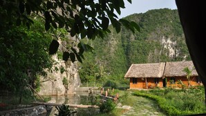 A rustic thatched-roof house by a calm river, surrounded by lush greenery and a rocky cliff. - Trang An Valley Bungalow (Tay Hoa Lu)