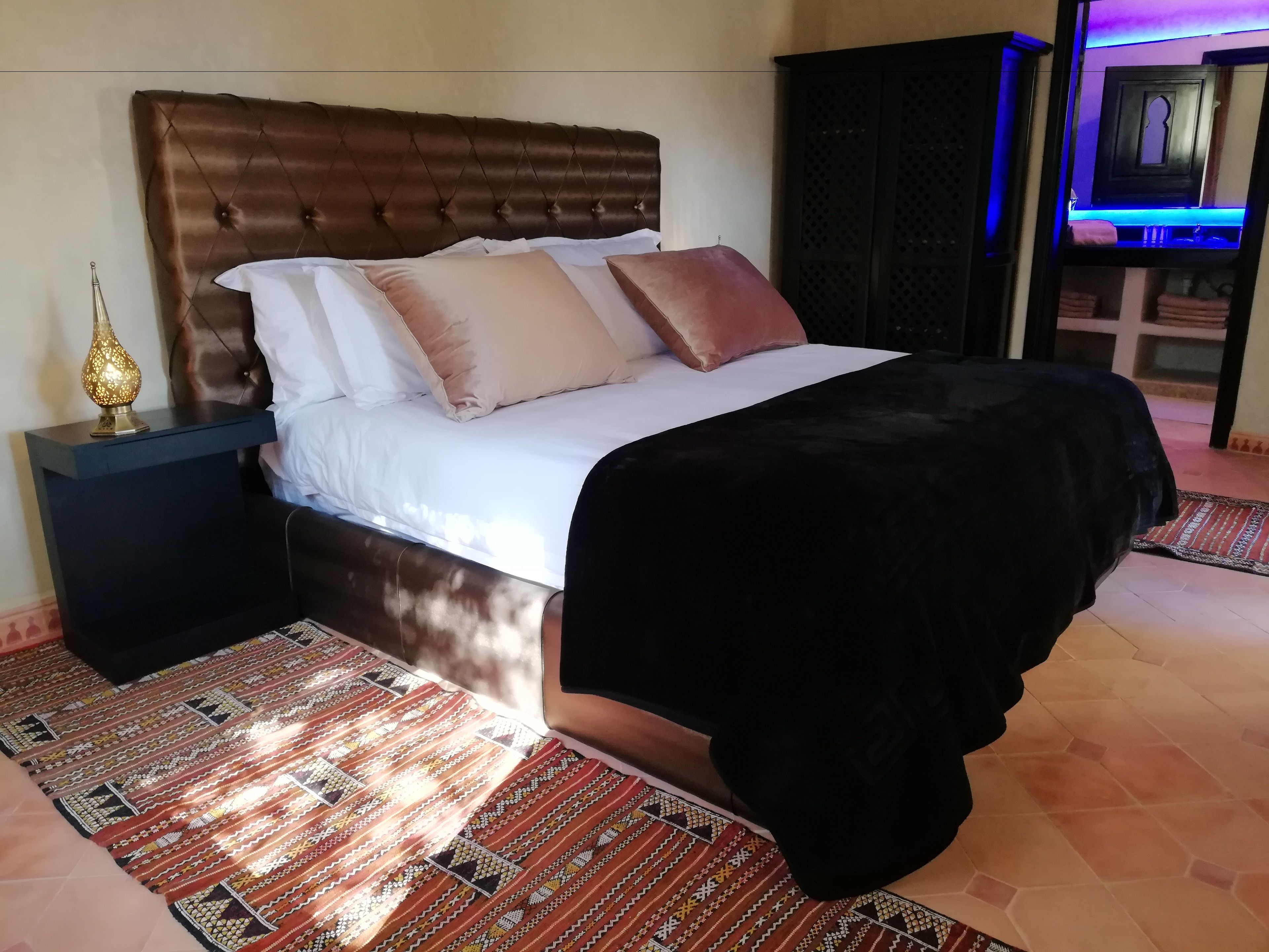 Una cama con cabecera capitonada, ropa de cama blanca y una manta de color negro. Hay una alfombra estampada en el suelo y una mesita de noche con una lámpara al lado de la cama.