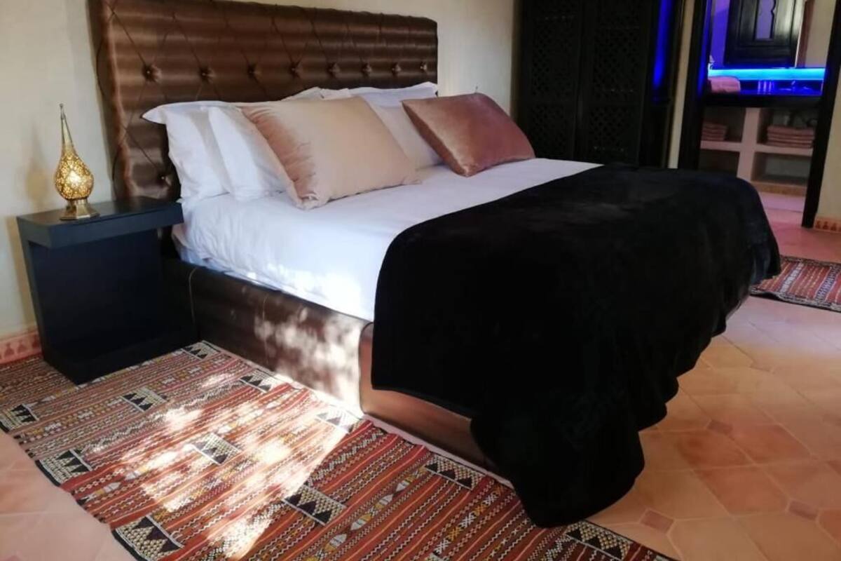 A bedroom with a tufted headboard, a patterned rug, a bedside table with a lamp, and a black bedspread.