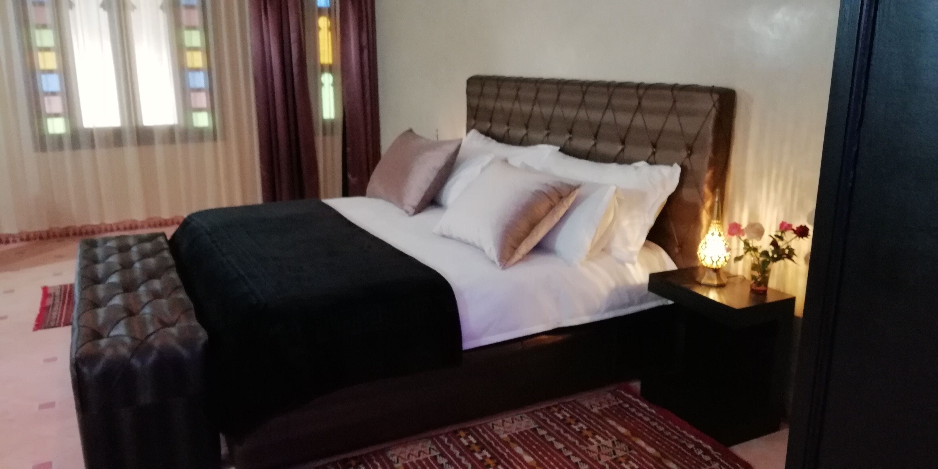 A neatly made bed with a dark headboard and white bedding, a bedside table with a lamp, and a patterned rug on the floor.