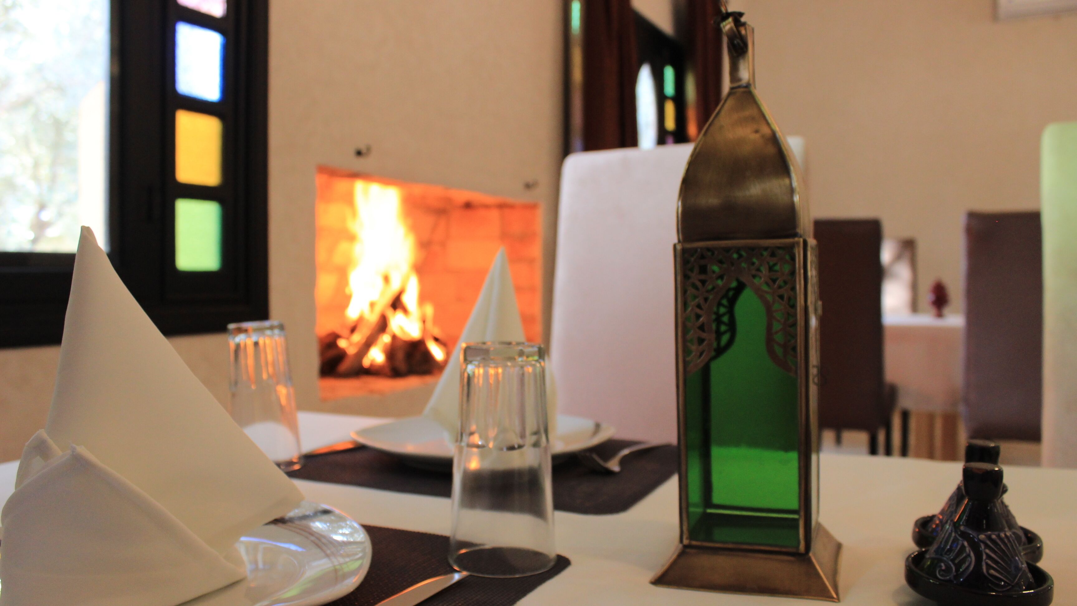 A table set with a glass, a candle, a napkin cone, a glass candle holder, and a decorative bottle. In the background, there is a fireplace with a visible fire.