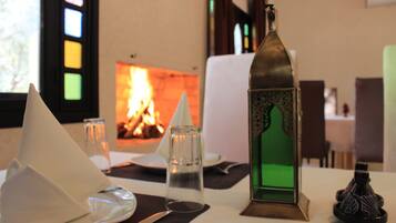 A table set with a glass, a candle, a napkin cone, a glass candle holder, and a decorative bottle. In the background, there is a fireplace with a visible fire.