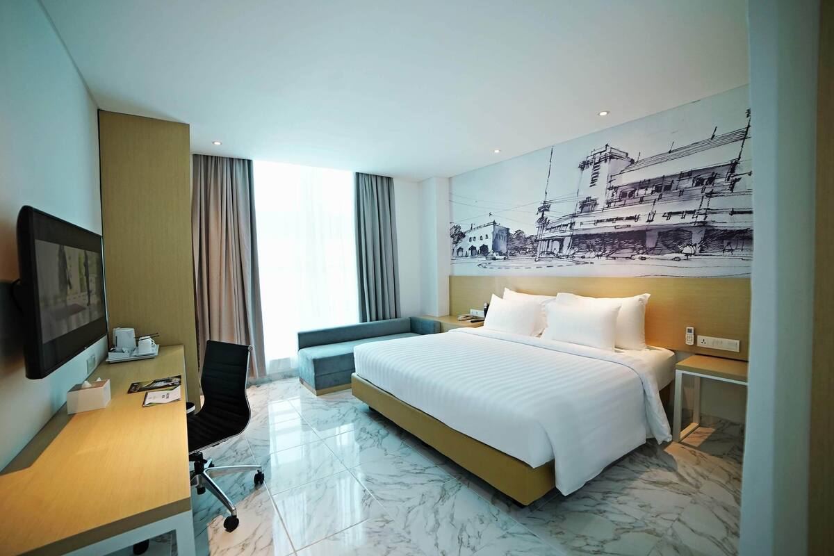 Ein Hotelzimmer mit einem großen Bett, einem Schreibtisch mit Computer, einem Sessel und einem Fernseher.