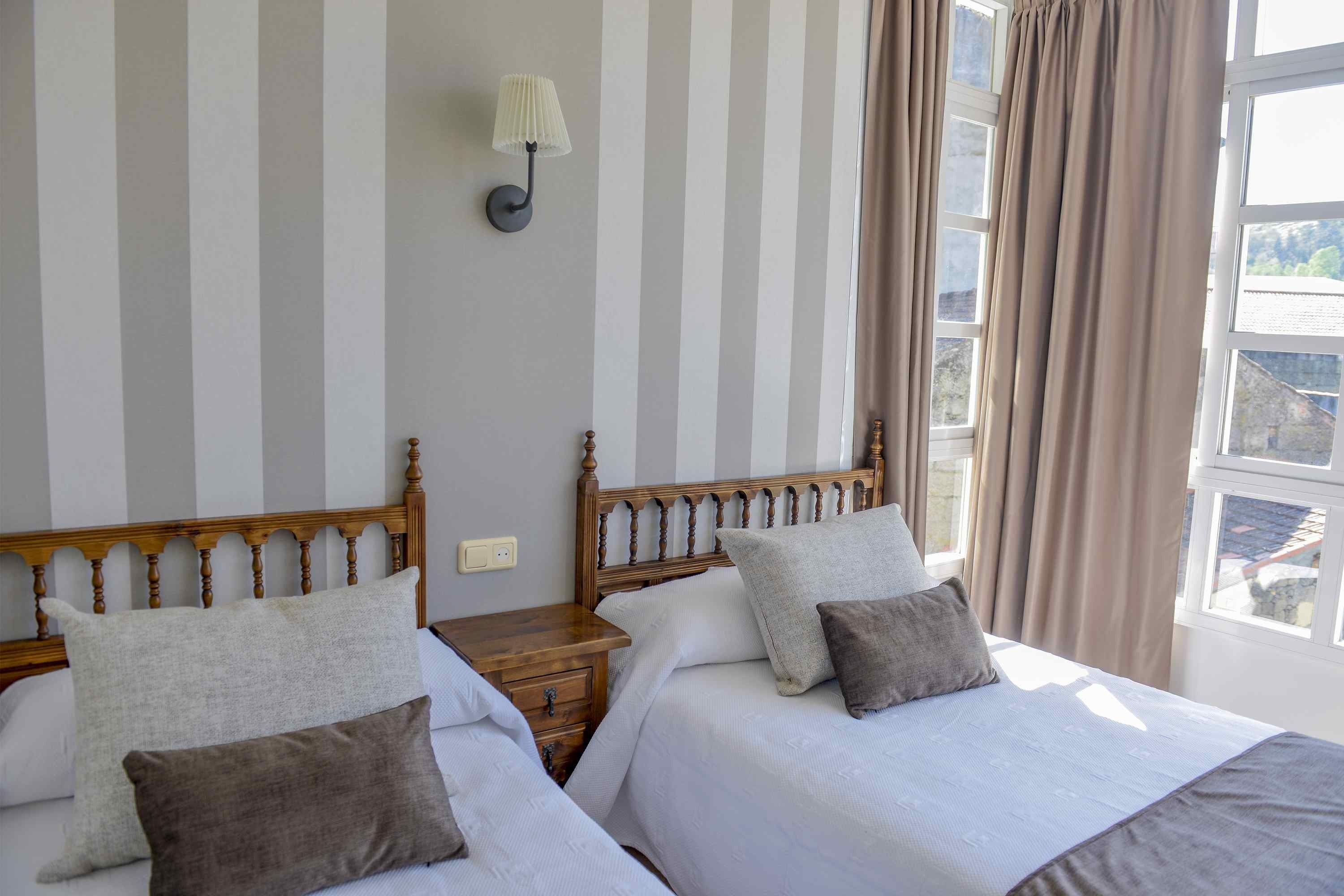 A double bed with two pillows, a nightstand, a wall-mounted lamp, and a window with curtains.