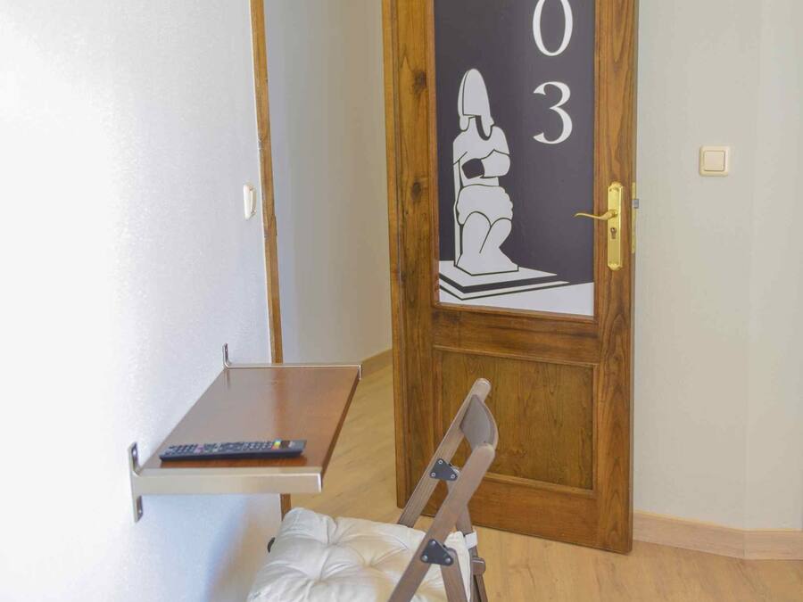 Una habitación con una puerta de madera que indica '203', una mesita con un control remoto y una silla plegable.