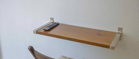 Una silla con asiento acolchado y respaldo de madera, ubicada debajo de un estante montado en la pared donde hay un control remoto.