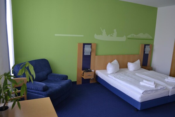 A hotel room with a bed, a blue armchair, a wooden nightstand, and a wall mural.