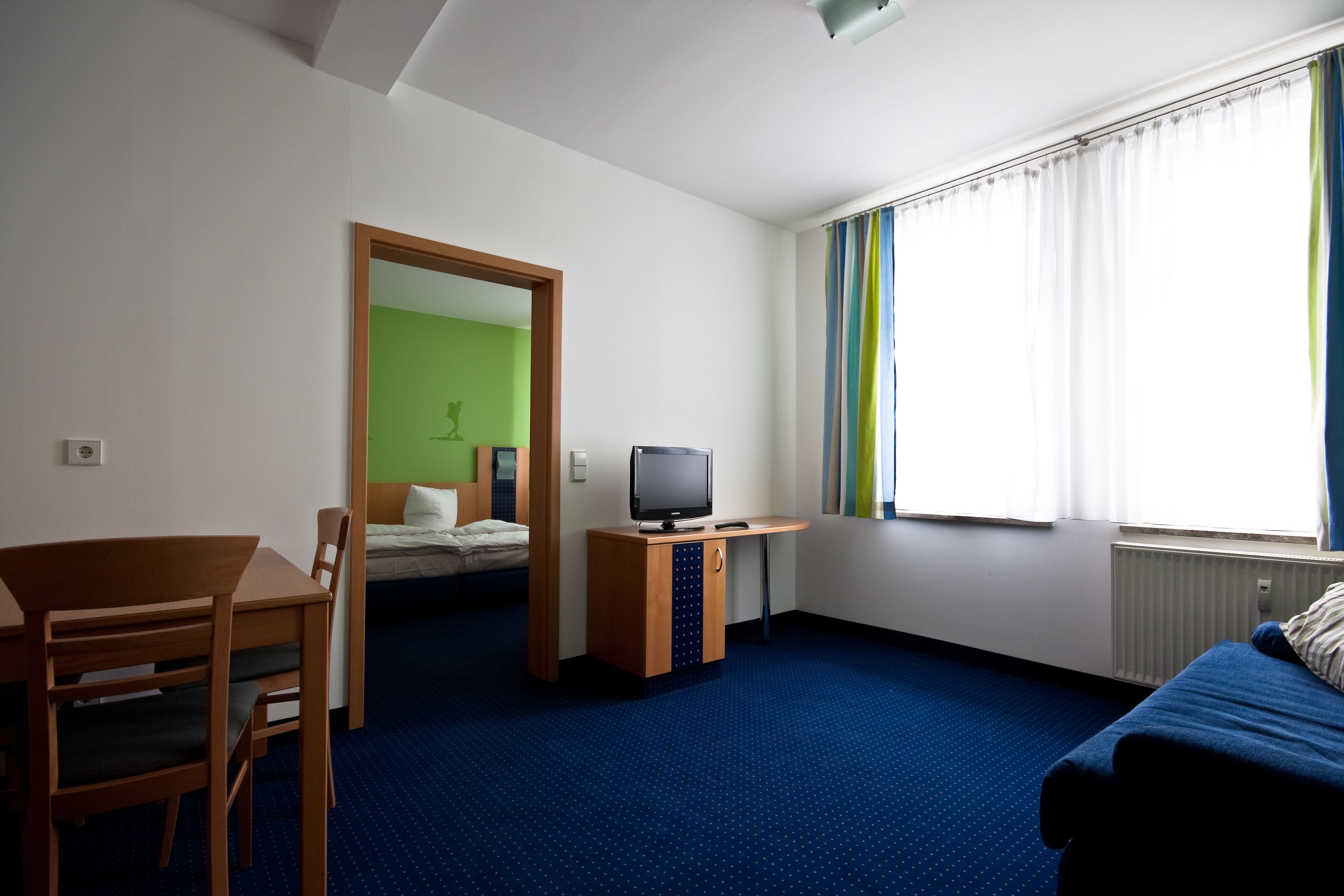 A hotel room with a bed, a desk with a television, a dining table, and a window with curtains.