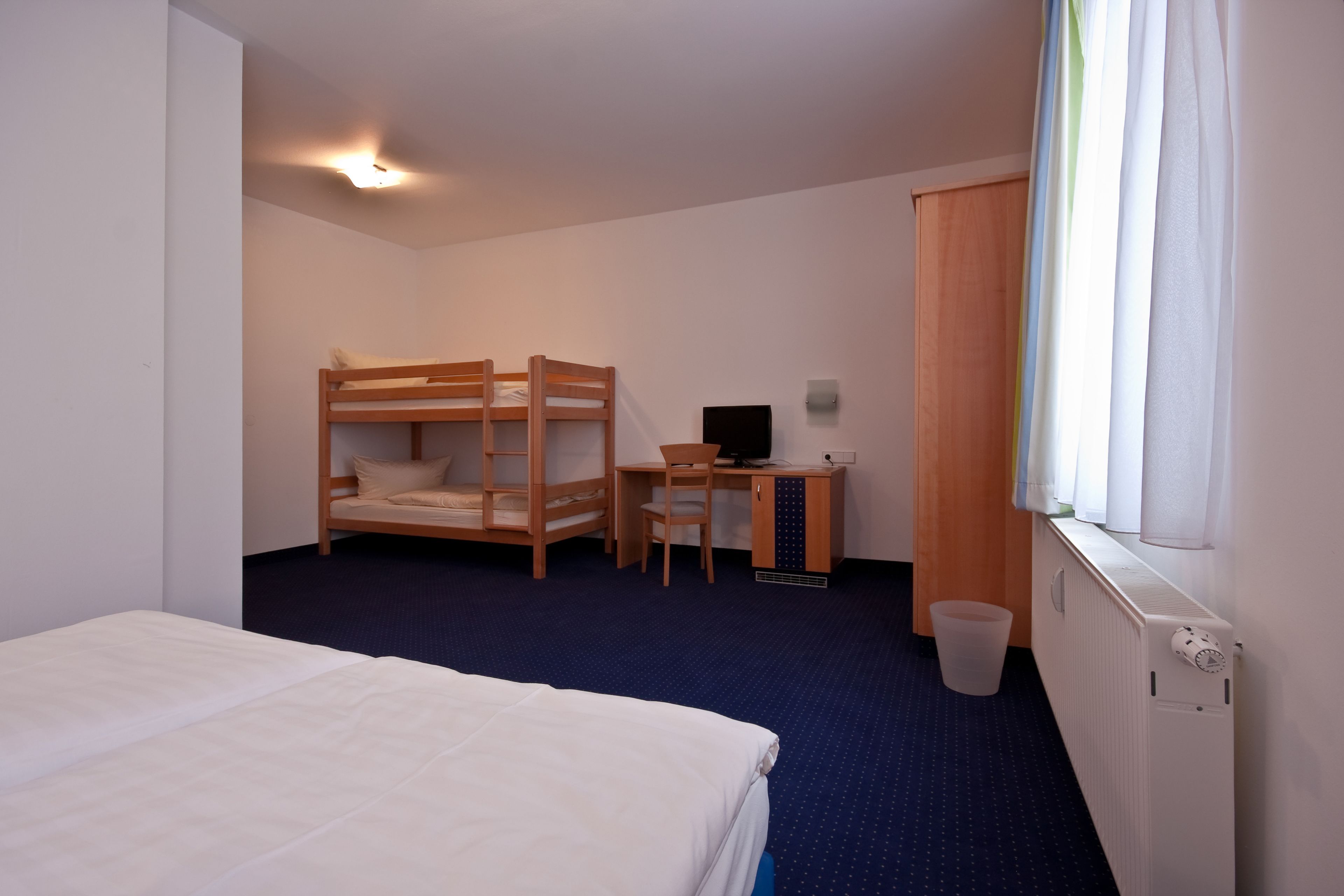 A hotel room with a bunk bed, a desk with a chair, a computer, a radiator, and a window with curtains.