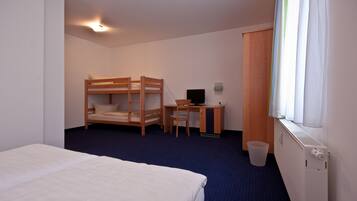 A hotel room with a bunk bed, a desk with a chair, a computer, a radiator, and a window with curtains.
