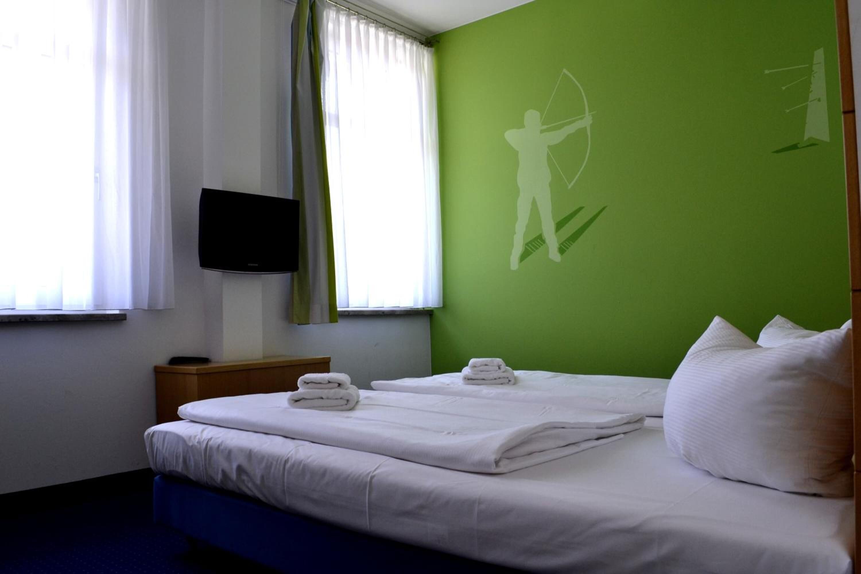 Habitación de hotel con una cama, un televisor montado en la pared y una pared verde con la silueta de una persona sosteniendo un arco.