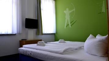 Habitación de hotel con una cama, un televisor montado en la pared y una pared verde con la silueta de una persona sosteniendo un arco.