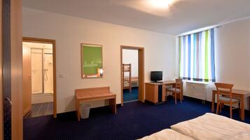 A hotel room with a bed, a desk with a chair, a TV, a bench, and a shower area.