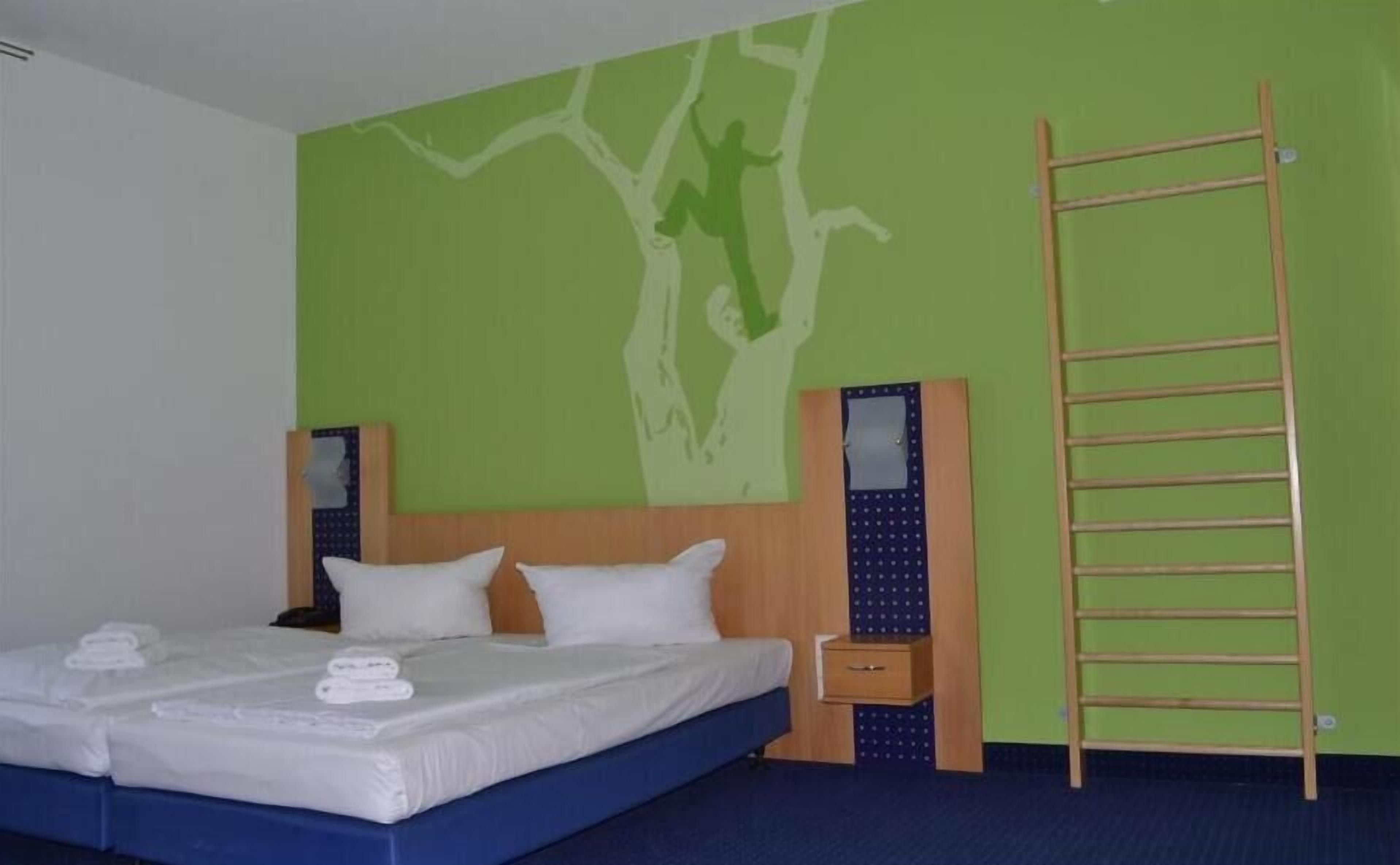 A hotel room with a bed, a wooden ladder, and a green wall with a tree mural.