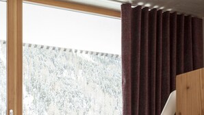 A bed with a white mattress and a floral cushion, a bedside lamp, and a view of a forest through a window with curtains. - Langtauferer Hof (Curon Venosta)