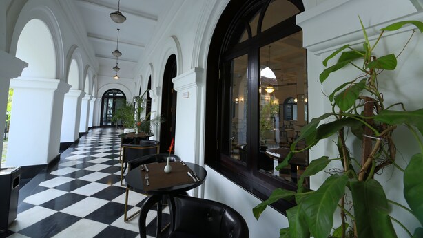 A corridor with black and white checkered flooring, white columns, and tables with chairs. - Alpha Old Court House (Kochi)