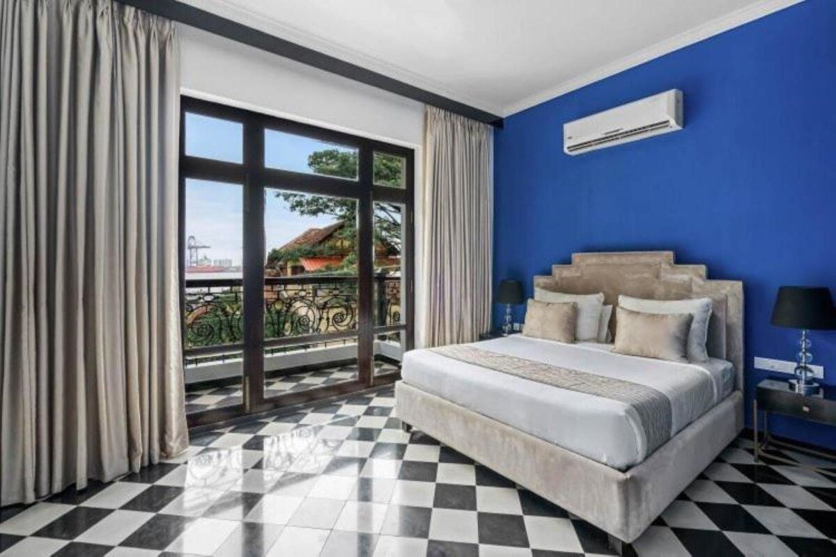 A hotel room with a checkered floor, a large bed with multiple pillows, a nightstand with a lamp, and a view of a balcony with a railing.