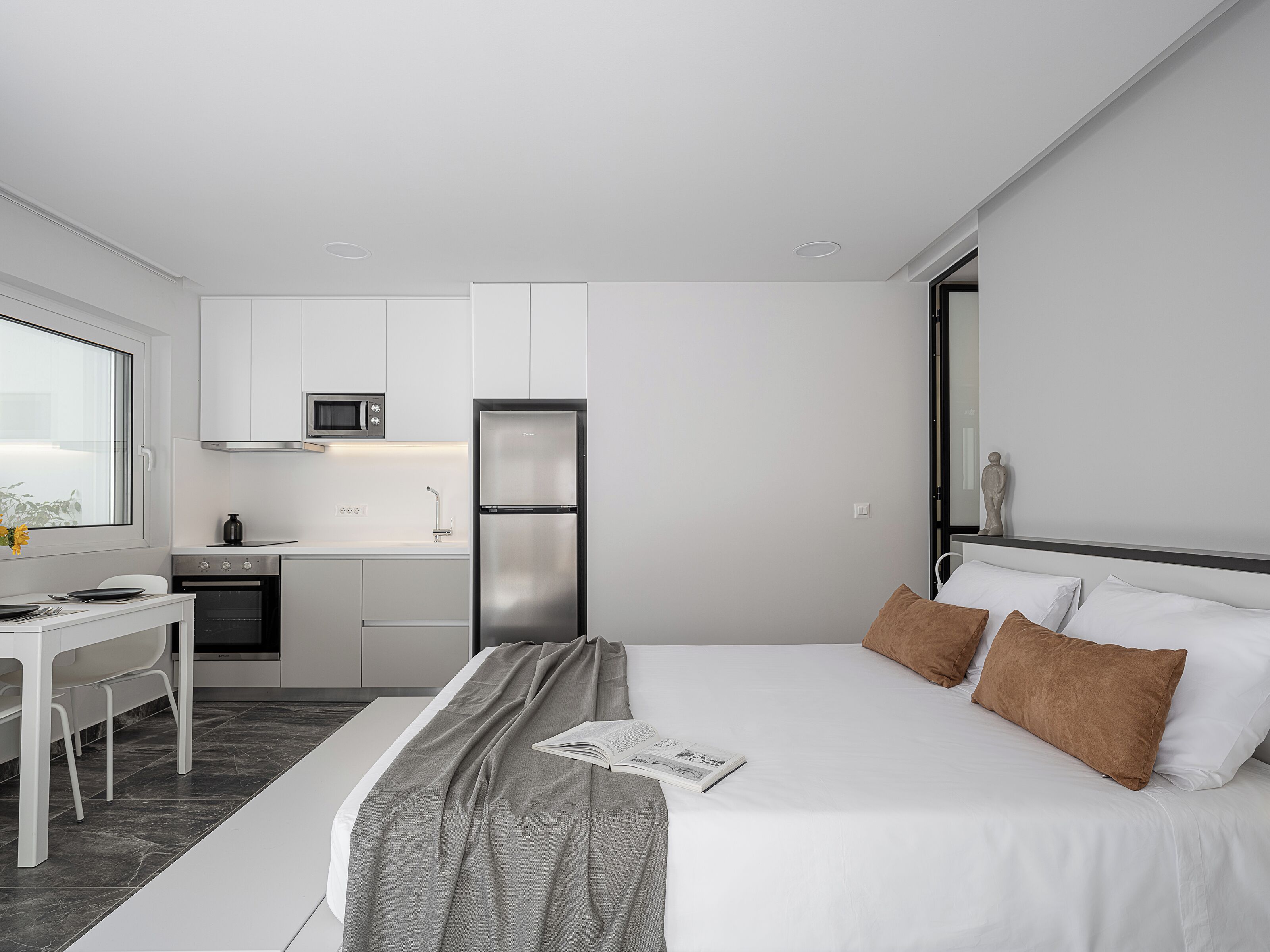 A modern hotel room with a bed, bedside table, and a small kitchenette.