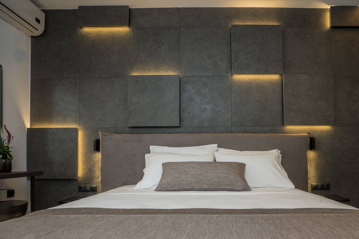 A modern bedroom with a large bed, a headboard with integrated lighting, a bedside table, and a wall-mounted air conditioning unit.