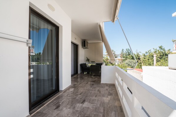 Ein Balkon mit Glastür, eine Essecke mit Tisch und Stühlen sowie ein Blick auf Grünflächen und Gebäude im Hintergrund.