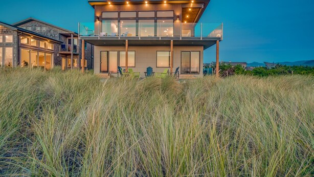 A modern two-story house with large windows and a balcony, surrounded by tall grass.