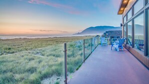 A balcony with a view of the beach and ocean, featuring outdoor seating and a glass railing.